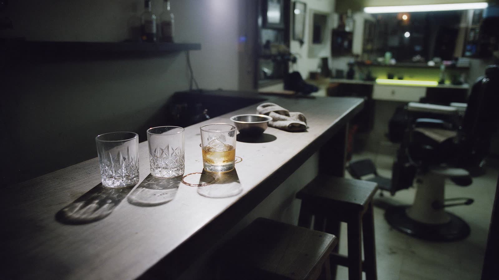 La barra del local con vasos tallados, uno servido con el hielo a medio derretir, y los sillones de barbero al fondo.
